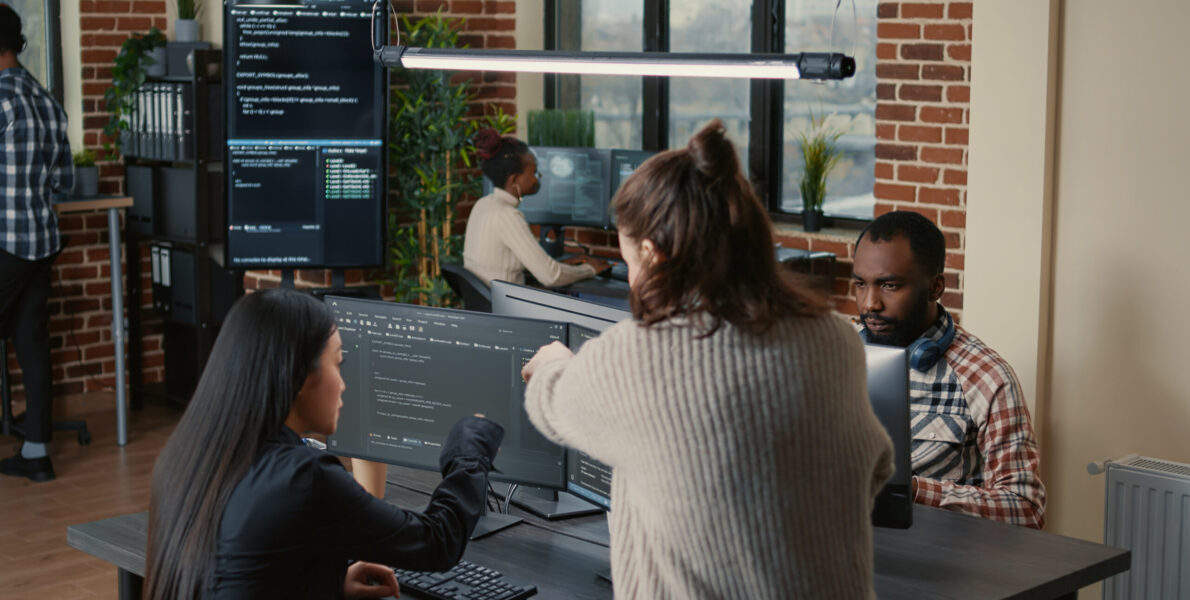 Software programer pointing pencil at source code on computer screen explaining algorithm to coworker standing next to desk. Programmers discussing online cloud computing with team.